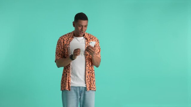 Young dark-skinned man looking at cellphone's screen while expecting for result of online betting. Winning man rejoicing, showing yes gesture, expressing success and victory, studio shot