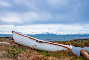 White boat moored on grassy field at seaside. Nautical vessel on land with snowcapped mountains in background. View of beautiful seascape against cloudy sky during winter.