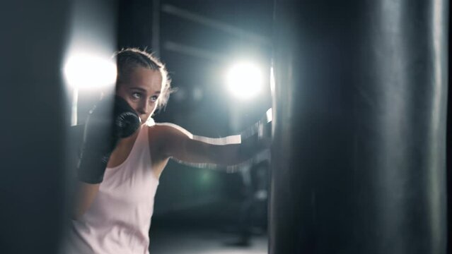 A muscular alone woman is hits punching bag in the gym. Sportswoman is boxing in dark hall. Self confident woman is training indoors. Maximum focus and strength. Concept of health and sports.