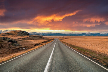 View of empty vanishing street amidst volcanic landscape. Scenic view of mountain range against dramatic sky. Road markings on diminishing road during sunset.