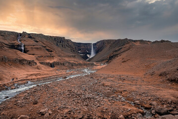 Scenic view of Litlanesfoss and Hengifoss in Eastfjords. Flowing stream from cliffs amidst dramatic landscape against cloudy sky. Scenic view of famous tourist attraction during sunset.