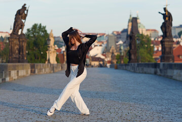Young woman in casual stylish clothes dancing on the stone bridge of old town in a Prague. Active...