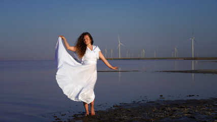 Graceful curly-haired girl in a long white dress on the background of wind power plants. pregnant girl in the sea. healthy ecological future