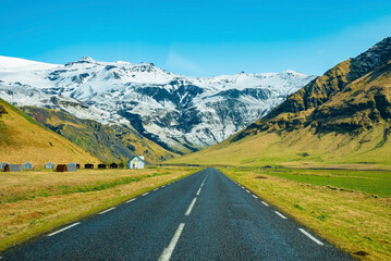 Diminishing empty road leading towards snowcapped mountain. Road markings on street amidst grassy landscape. Scenic view of highway with houses on roadside against blue sky.