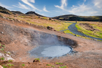 High angle view of geothermal mud spot against mountains. View of road amidst landscape in background. View of steaming fumarole against sky in Hveragerdi valley.