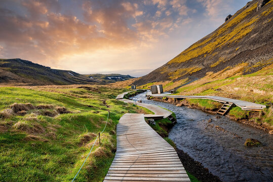 Beautiful view of wooden boardwalk by hot river stream. Natural geothermal bath in Reykjadalur Valley against cloudy sky. Grassy landscape by mountains in Hveragerdi during sunset.