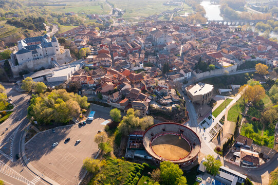 Aerial View Of Small Spanish Township Of Simancas Overlooking Brownish Tiled Roofs Of Residential Buildings, Modern Bullring And Medieval Fortified Castle In Background On Sunny Spring Day