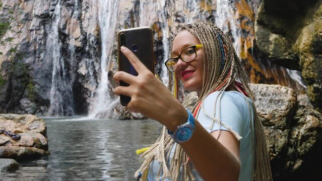 Cute female in yellow glasses use smartphone take selfie on waterfall background. Take picture of yourself. Woman smiles, water pond surface with stone
