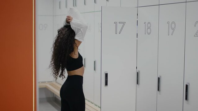 Young lady athlete taking off her t-shirt, getting ready for fitness workout, standing in front of locker at gym