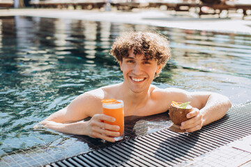 Curly positive guy holds cocktails and enjoys spending time in the pool