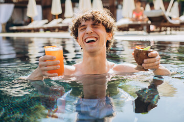 Curly positive guy holds cocktails and enjoys spending time in the pool