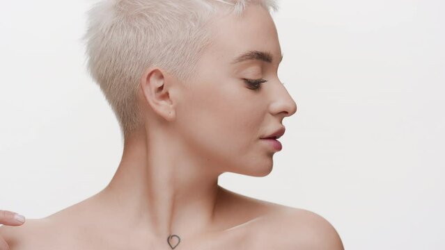Gorgeous young slim Caucasian woman with light hair strokes her collarbones turning her head on white background | Body moisturizing concept