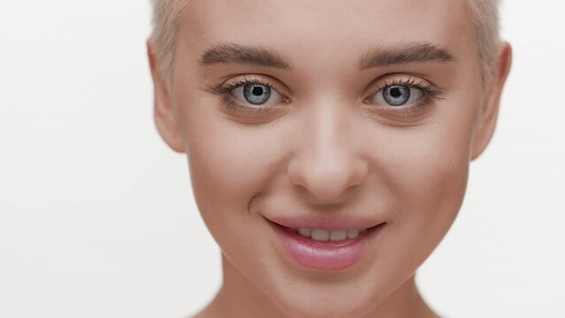 Big close-up shot of beautiful slim young blond Caucasian woman opens her eyes and smiles wide for the camera looking at it on white background | Face care concept
