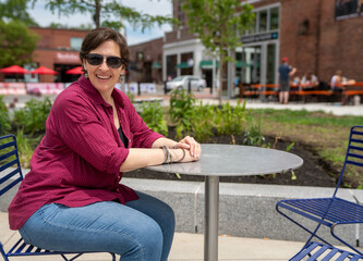 Fototapeta premium Happy, Mature woman sitting alone at an outdoor table wearing sunglasses.