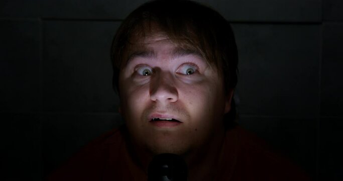 There are power outages in the house, so a young man is sitting in the dark with a terrified look and illuminates his face with flashlight, front view, close up