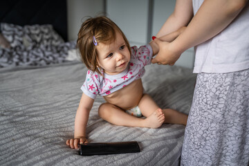 Fototapeta premium One small caucasian girl toddler sitting on the bed at home watching tv while her mother is changing her clothes real people growing up copy space