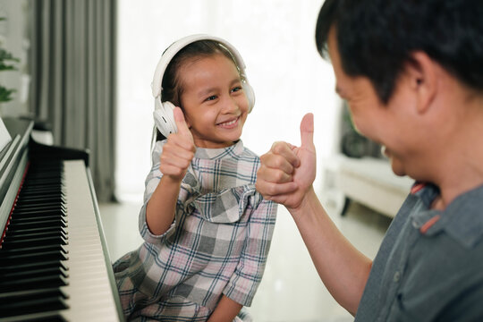 Asian daughter and father showing thumb and smiling together while playing piano at home, concept of love, bonding, relation, education, music, skill, mental health, parent and child in family life.