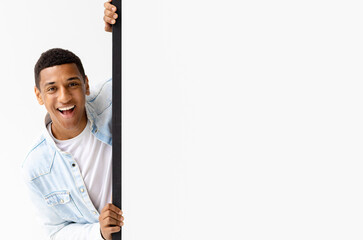 Young African American man peeking out from empty white board, looks at the camera and smiles. Copy...