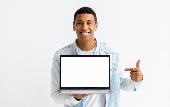 Portrait Of Young African American Man With Laptop On White Background. Male Points Her Finger At A Blank Laptop Screen, Looking At The Camera And Smiling