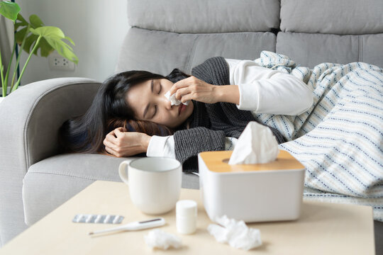 Upset young Asian woman sleeping on couch covered with blanket freezing blowing running nose got fever caught cold sneezing in tissue, sick female having influenza symptoms sleep at home. Flu concept