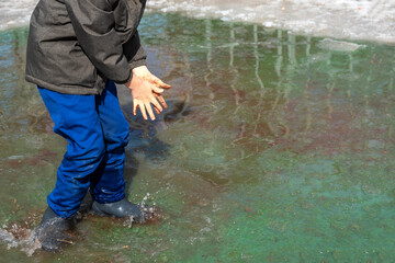 Joyfull Child in rubber boots kneading dirt with hands in muddy puddle. Environmental pollution. Child boy of 6-7 year old walking and playingin wellies in polluted water