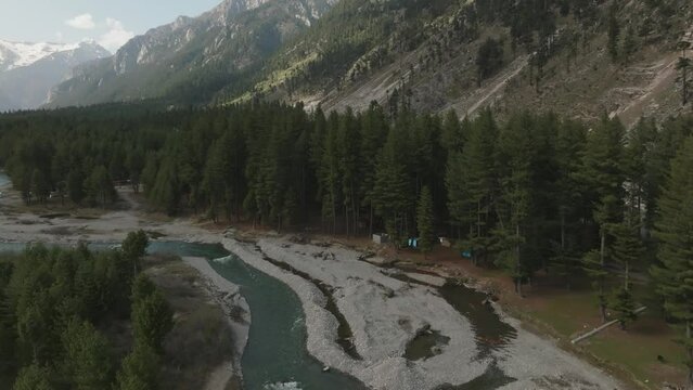 Aerial, flying in top of a river in the middle of a pine forest in the Himalayas, mountains with snow in the  back of the forest, drone goes down slowly. Kumrat, KPK, Pakistan