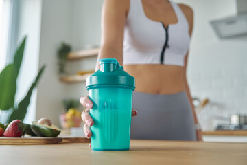 Close-up of woman in sports clothing holding shaker with protein cocktail at the kitchen