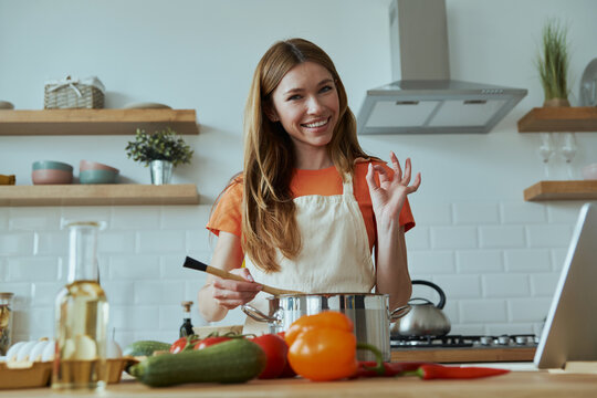 Confident young woman cooking soup and gesturing while standing at the domestic kitchen