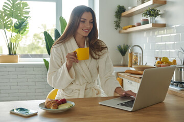 Beautiful woman in bathrobe enjoying breakfast and using laptop while spending time in the kitchen