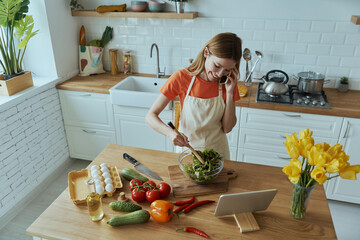 Top view of cheerful young woman talking on mobile phone while cooking at the kitchen