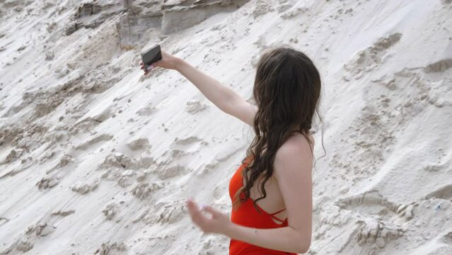 Woman brunette in red swimsuit with bare back makes selfie on beach sandy slope. Lady poses taking picture on phone. Summer tropical vacation