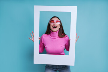 Excited young woman looking through a picture frame while standing against blue background