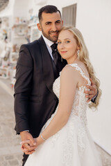 Portrait of young blonde bride in white dress and brunet groom in suit smiling and embracing while walking down street in Italy closeup, urban background. Beautiful and romantic wedding, happy couple