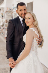 Naklejka premium Portrait of young blonde bride in white dress and brunet groom in suit smiling and embracing while walking down street in Italy closeup, urban background. Beautiful and romantic wedding, happy couple