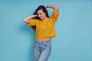 Beautiful young woman holding hands in hair and smiling while standing against blue background