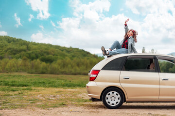 Funny teen girl is sitting on the roof of a car. Mountains and sky are in the background. Copy space. The concept of freedom and car trip
