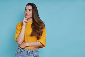 Thoughtful young woman holding hand on chin while standing against blue background