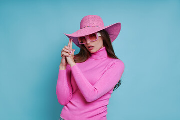 Attractive woman in pink hat gesturing gun while standing against blue background
