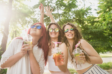 Three young women in fashionable clothes and bright sunglasses walk around the city in summer and drink refreshing non-alcoholic cocktails from disposable plastic cups. Women's friendship and walks in