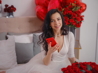 young woman in white dres is sitting on bed and chatting on smartphone with boyfriend. Anniversary, happy birthday, wedding , love day, women's day, Valentine day and positive emotions