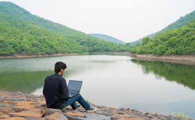 Fototapeta premium Young man working on a laptop and sitting in the nature, Workation and Digital Nomad concept.
