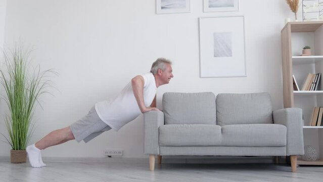 Sofa workout. Home fitness. Morning training. Body sculpt. Strong athletic middle-aged man doing push-up exercise on couch in light living room interior.
