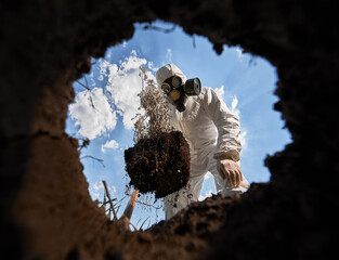 View from inside pit of ecologist digging pit by shovel and planting tree, wearing gas mask and...
