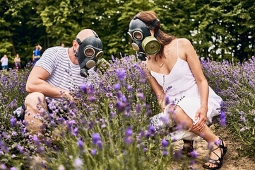 Couple crouching in lavender field wearing gas masks. Blooming purple flowers blurred on...