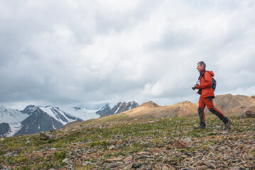 Obraz premium Man in red with camera walks with view to large snowy mountain range with sharp tops under gray cloudy sky. Dramatic landscape with tourist in high mountains. Snow pointy peaks at changeable weather.