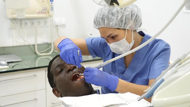 Asian woman orthodontist using tools and doing oral care inspection on african-american man patient to cure toothache.