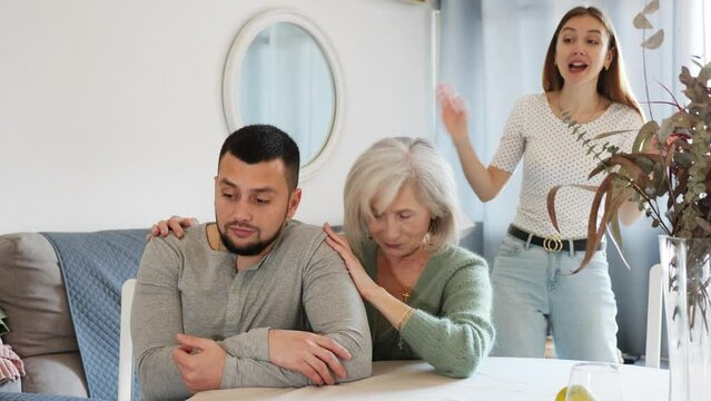 Portrait of displeased young adult man sitting at home ignoring his wife berating him, while her mother embracing and soothing him