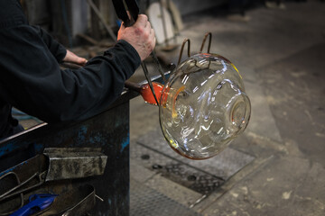 Man twists a blowpipe making a glass object and forms a neck to the hot glass object with pliers in the glassworks
