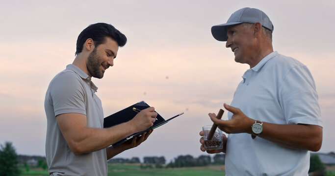 Two men talking golf course outside. Golfers analyze game result at sunset field
