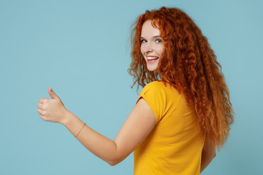 Back rear view young smiling fun happy redhead woman 20s wearing yellow t-shirt showing thumb up like gesture isolated on plain light pastel blue background studio portrait. People lifestyle concept.